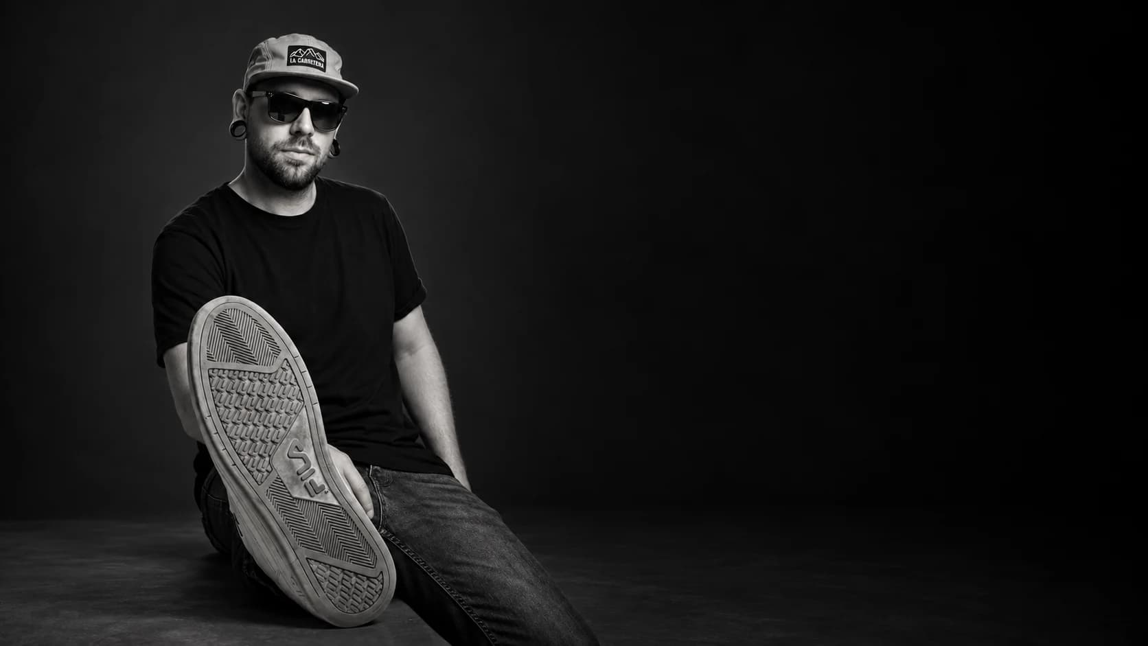 Edu García Devesa, de edugdstudio, sentado de tres cuartos sobre un bloque, con gafas de sol y gorra, la suela de la zapatilla hacia la cámara